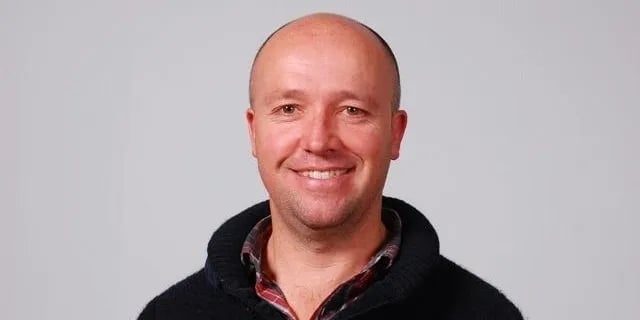 Headshot of a smiling bald man wearing a black hoodie over a red plaid shirt against a light gray background